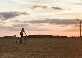 Cyclist rides through field with wind turbine at sunset, symbolizing renewable energy.